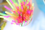 Colorful plastic straws arranged in a container, viewed from above. The straws are in bright neon shades including pink, yellow, green, and orange. The background is blurred and appears to be in soft, light colors.