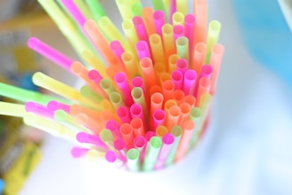 Colorful plastic straws arranged in a container, viewed from above. The straws are in bright neon shades including pink, yellow, green, and orange. The background is blurred and appears to be in soft, light colors.