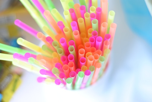 Colorful plastic straws arranged in a container, viewed from above. The straws are in bright neon shades including pink, yellow, green, and orange. The background is blurred and appears to be in soft, light colors.
