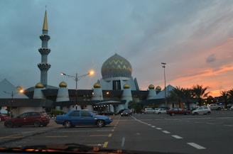 A large mosque with a prominent dome and tall minaret, featuring geometric patterns and gold detailing. The foreground has a parking lot with several cars. The sky is cloudy with a vibrant sunset creating a mix of warm hues.