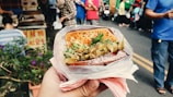 A hand holds a plastic-wrapped sandwich filled with vegetables and herbs, with a grilled top. The background features a busy street market with people and stalls.