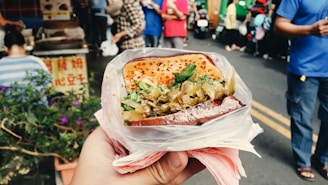A hand holds a plastic-wrapped sandwich filled with vegetables and herbs, with a grilled top. The background features a busy street market with people and stalls.