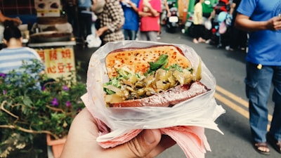 A hand holds a plastic-wrapped sandwich filled with vegetables and herbs, with a grilled top. The background features a busy street market with people and stalls.
