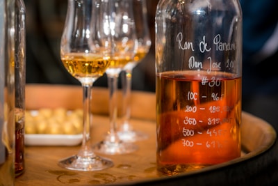 A close-up of a bottle filled with amber liquid, presumably rum, placed on a wooden barrel. The bottle is inscribed with writing indicating measurements. Several wine glasses containing a similar colored liquid are also visible, and there is a plate with light-colored snacks in the background.