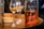 A close-up of a bottle filled with amber liquid, presumably rum, placed on a wooden barrel. The bottle is inscribed with writing indicating measurements. Several wine glasses containing a similar colored liquid are also visible, and there is a plate with light-colored snacks in the background.