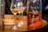 A close-up of a bottle filled with amber liquid, presumably rum, placed on a wooden barrel. The bottle is inscribed with writing indicating measurements. Several wine glasses containing a similar colored liquid are also visible, and there is a plate with light-colored snacks in the background.
