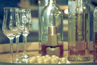 A collection of glassware featuring three elegant wine glasses with tall stems and clear, reflective surfaces is situated prominently. In the background, several bottles containing a rose-colored liquid are placed. There are also shiny, golden spherical objects scattered on a wooden surface.