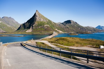 A picturesque landscape featuring a winding road leading towards a triangular mountain peak. The scene is surrounded by clear blue water, with smaller rocky islands scattered around. The landscape is lush with green vegetation and rugged mountains in the distance, all set under a clear blue sky.