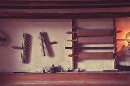 An array of vintage kitchen tools is displayed on a wall, including wooden rolling pins and spiked rollers. The tools are mounted on a textured wall, creating a rustic feel. Soft lighting casts shadows, enhancing the antique ambiance.