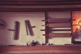 An array of vintage kitchen tools is displayed on a wall, including wooden rolling pins and spiked rollers. The tools are mounted on a textured wall, creating a rustic feel. Soft lighting casts shadows, enhancing the antique ambiance.