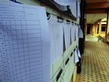 Several sheets of paper with printed tables are clipped to a bulletin board in a corridor. The papers appear to contain data or results, indicated by numerical values and columns.