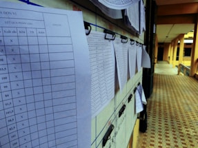 Several sheets of paper with printed tables are clipped to a bulletin board in a corridor. The papers appear to contain data or results, indicated by numerical values and columns.