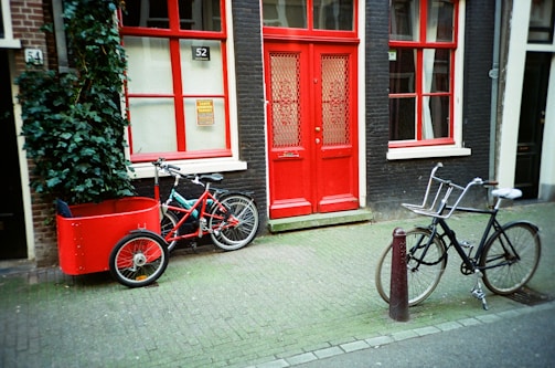 A street scene featuring two bicycles parked in front of a building with bright red doors and windows. One bicycle is fitted with a red cargo box, while the other is a traditional black bicycle. The building facade is dark, with red accents and a small number '52' displayed above the door. A green leafy plant climbs up the wall next to the cargo bicycle.