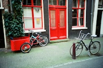A street scene featuring two bicycles parked in front of a building with bright red doors and windows. One bicycle is fitted with a red cargo box, while the other is a traditional black bicycle. The building facade is dark, with red accents and a small number '52' displayed above the door. A green leafy plant climbs up the wall next to the cargo bicycle.
