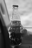 A hand is holding a classic 8 fl oz Coca-Cola glass bottle, which has a distinctive contour shape and a cap secured at the top. The background is slightly blurred, suggesting motion or focus on the bottle.