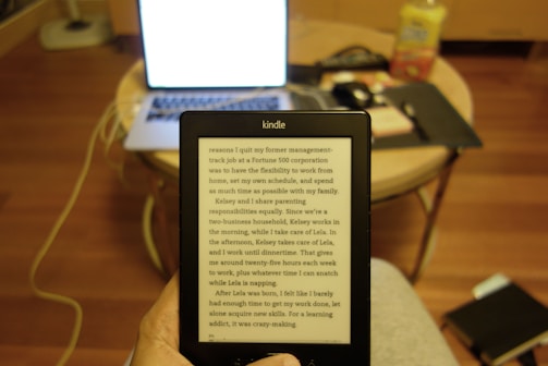 A Kindle displaying text is held by a person in front of a laptop with a bright screen. The setting appears to be a domestic environment. The laptop is on a wooden round table which also holds other items like a phone, a notebook, and a bottle. A wooden floor is visible, and the furniture creates a cozy, home-office atmosphere.
