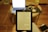 A Kindle displaying text is held by a person in front of a laptop with a bright screen. The setting appears to be a domestic environment. The laptop is on a wooden round table which also holds other items like a phone, a notebook, and a bottle. A wooden floor is visible, and the furniture creates a cozy, home-office atmosphere.