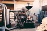 An industrial coffee roasting machine with a person working on its side, surrounded by bags of coffee beans and blue plastic bins in a rustic interior setting with exposed beams and pipes.