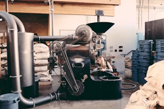 An industrial coffee roasting machine with a person working on its side, surrounded by bags of coffee beans and blue plastic bins in a rustic interior setting with exposed beams and pipes.