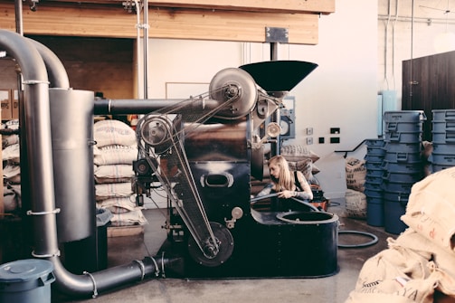 An industrial coffee roasting machine with a person working on its side, surrounded by bags of coffee beans and blue plastic bins in a rustic interior setting with exposed beams and pipes.