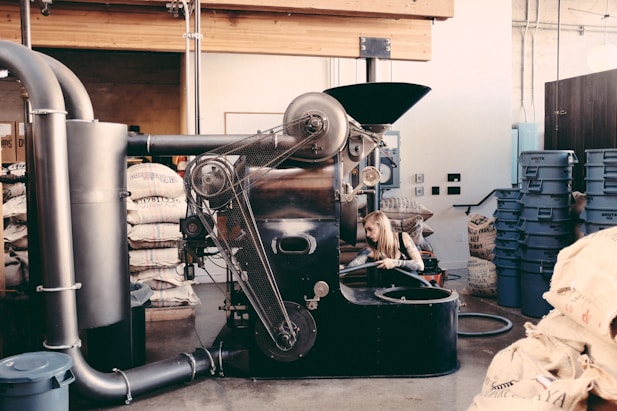 An industrial coffee roasting machine with a person working on its side, surrounded by bags of coffee beans and blue plastic bins in a rustic interior setting with exposed beams and pipes.