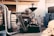 An industrial coffee roasting machine with a person working on its side, surrounded by bags of coffee beans and blue plastic bins in a rustic interior setting with exposed beams and pipes.