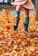 A person wearing a light dress, teal knee-high socks, and brown boots is walking through a carpet of dried autumn leaves. The leaves are scattered and kicked up in motion, suggesting a playful interaction with the falling foliage. The vibrant autumn colors of the leaves create a warm and lively atmosphere.