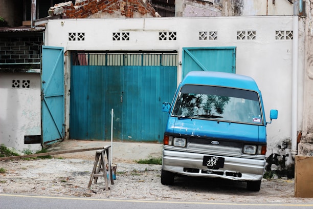 A blue van is parked slightly slanted in front of a worn, industrial-looking garage with a corrugated metal door. The garage and nearby buildings have visible signs of aging, with exposed bricks and peeling paint. There is also a small partially open blue door beside the vehicle. The scene is set in an urban environment, with a focus on rugged and utilitarian features.