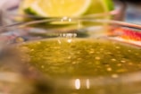 A close-up view of a bowl containing green salsa with visible seeds and a semi-thick consistency. In the background, a blurred lime has been cut into quarters, enhancing the fresh and zesty impression of the scene.