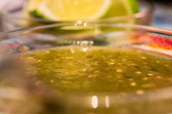 A close-up view of a bowl containing green salsa with visible seeds and a semi-thick consistency. In the background, a blurred lime has been cut into quarters, enhancing the fresh and zesty impression of the scene.