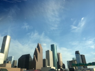 A city skyline featuring diverse skyscrapers against a clear blue sky. The buildings vary in design and height, with some having reflective glass surfaces. Traffic signs are visible in the foreground, indicating an urban setting.
