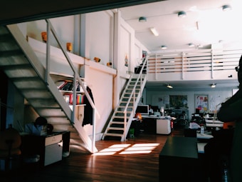 A spacious two-level workspace with wooden floors and white walls. An open staircase on the left and right leads to a mezzanine level with a railing. Below the mezzanine are desks with computers, shelves with books, and various office supplies. Natural light streams through windows, casting shadows on the floor. The workspace has a modern and organized appearance.