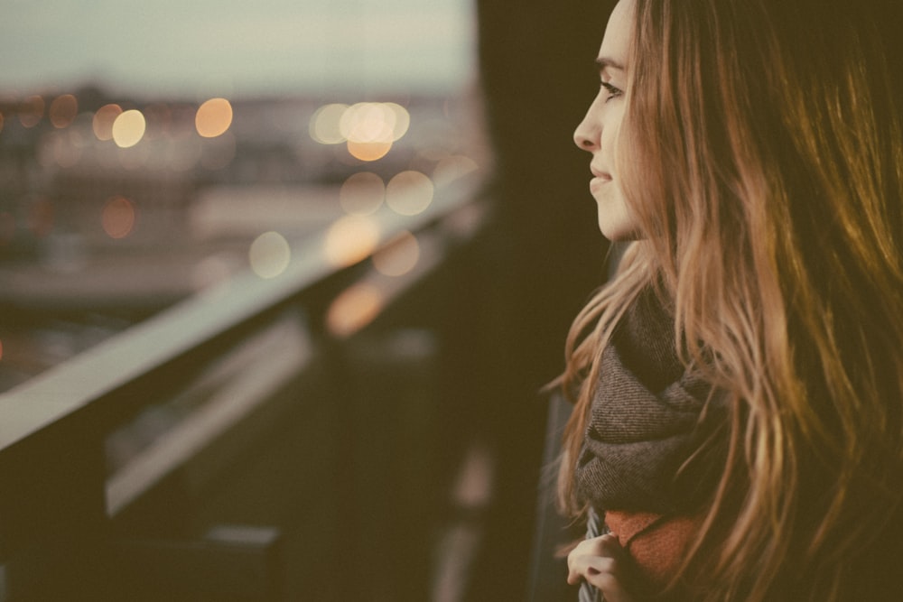 A young woman in a scarf smiling when looking off into the city from a balcony