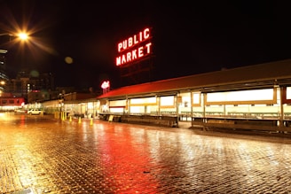 A nighttime view of a public market with bright, illuminated signage that reads 'PUBLIC MARKET' in red against a dark sky. The street is wet, reflecting the market lights, and appears to be paved with cobblestones. Several storefronts are visible, and there are a few parked vehicles and traffic cones along the side.