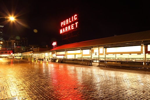 A nighttime view of a public market with bright, illuminated signage that reads 'PUBLIC MARKET' in red against a dark sky. The street is wet, reflecting the market lights, and appears to be paved with cobblestones. Several storefronts are visible, and there are a few parked vehicles and traffic cones along the side.
