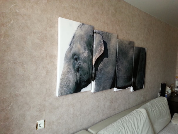 A wall-mounted, multi-panel canvas artwork depicting a close-up of an elephant. The image spans across four panels, each aligned to show a continuous perspective of the elephant&rsquo;s face and part of its body. The artwork hangs above a white leather couch against a textured beige wall. A power outlet is visible near the lower left corner.