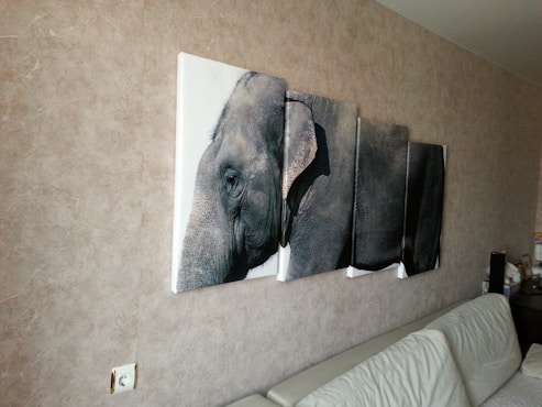 A wall-mounted, multi-panel canvas artwork depicting a close-up of an elephant. The image spans across four panels, each aligned to show a continuous perspective of the elephant&rsquo;s face and part of its body. The artwork hangs above a white leather couch against a textured beige wall. A power outlet is visible near the lower left corner.
