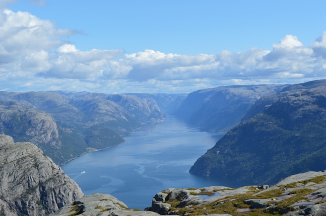 A stunning view of a deep blue fjord cutting through rugged, rocky cliffs and mountains, under a sky dotted with scattered clouds. The landscape is lush with greenery, particularly on the slopes of the mountains, contrasting with the rocky peaks.