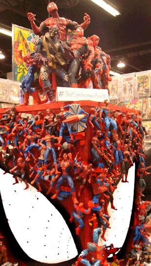 A large display of numerous Spider-Man figurines arranged densely on a booth structure, resembling a giant Spider-Man head. Some other comic characters are interspersed among the Spider-Man figures. Comic books can be seen in the background.