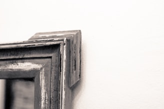 Close-up of the corner of a weathered and distressed wooden picture frame mounted on a white wall. The frame appears to have a vintage or rustic look with visible wood grain and cracked paint.