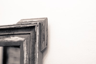 Close-up of the corner of a weathered and distressed wooden picture frame mounted on a white wall. The frame appears to have a vintage or rustic look with visible wood grain and cracked paint.