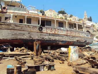 An old, weathered ship named 'Lazy Days' is docked on a beach or shipyard, surrounded by wooden planks, barrels, and debris. The ship's paint is peeling, revealing the wear and tear from exposure to the elements. Behind the ship, there are colorful buildings situated on a hillside under a clear blue sky.