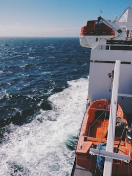 A lifeboat attached to the side of a ship with a view of the ocean and waves. The lifeboat is bright orange and secured to the ship's deck. The sea is dark blue with visible churning white waves, indicating movement or wind. The sky is clear with a hint of clouds on the horizon.