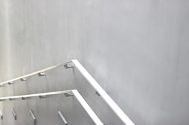 A minimalist staircase with sleek metal handrails is positioned against a plain gray wall. The design appears modern with clean lines and an industrial aesthetic.