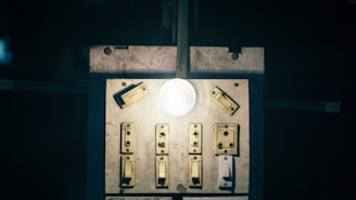 A dimly lit switchboard with multiple old-fashioned switches and a glowing light bulb in the center. The surface appears worn and aged, suggesting frequent use.
