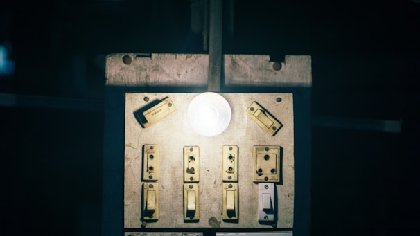 A dimly lit switchboard with multiple old-fashioned switches and a glowing light bulb in the center. The surface appears worn and aged, suggesting frequent use.