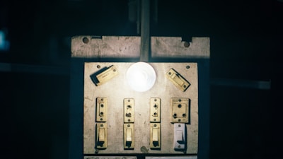 A dimly lit switchboard with multiple old-fashioned switches and a glowing light bulb in the center. The surface appears worn and aged, suggesting frequent use.