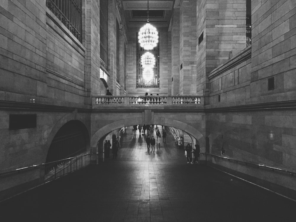 people walking on hallway of concrete building with turned on chandelier lights in grayscale photography