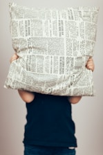 A person is holding a pillow covered with newspaper print patterns in front of their face. The person is wearing a dark blue top and is visible from the shoulders down. The background is plain and light-colored.