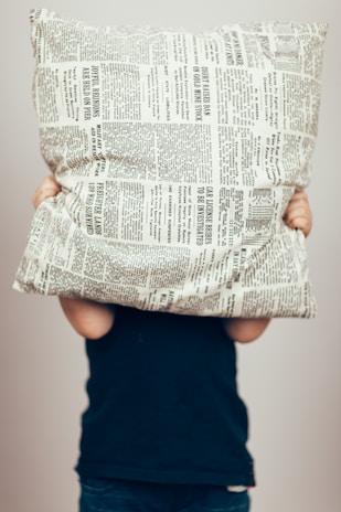 A person is holding a pillow covered with newspaper print patterns in front of their face. The person is wearing a dark blue top and is visible from the shoulders down. The background is plain and light-colored.